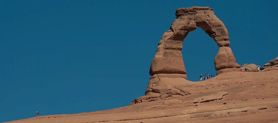 Delicate Arch rises above sloping sandstone, with small visitors beneath the opening providing scale.