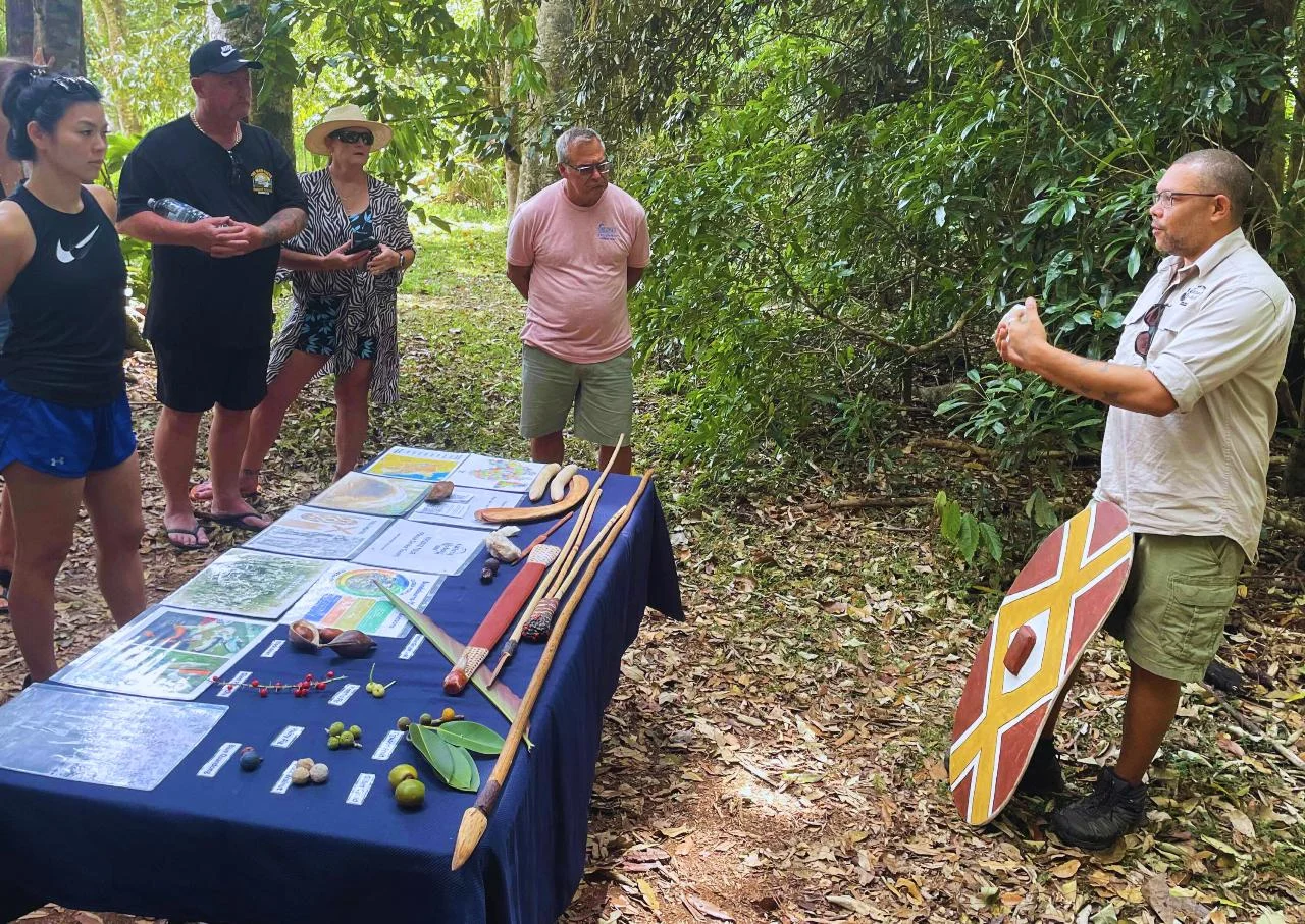 A cultural guide stands beside a table of mapped plants, foods and tools while visitors listen in the rainforest
