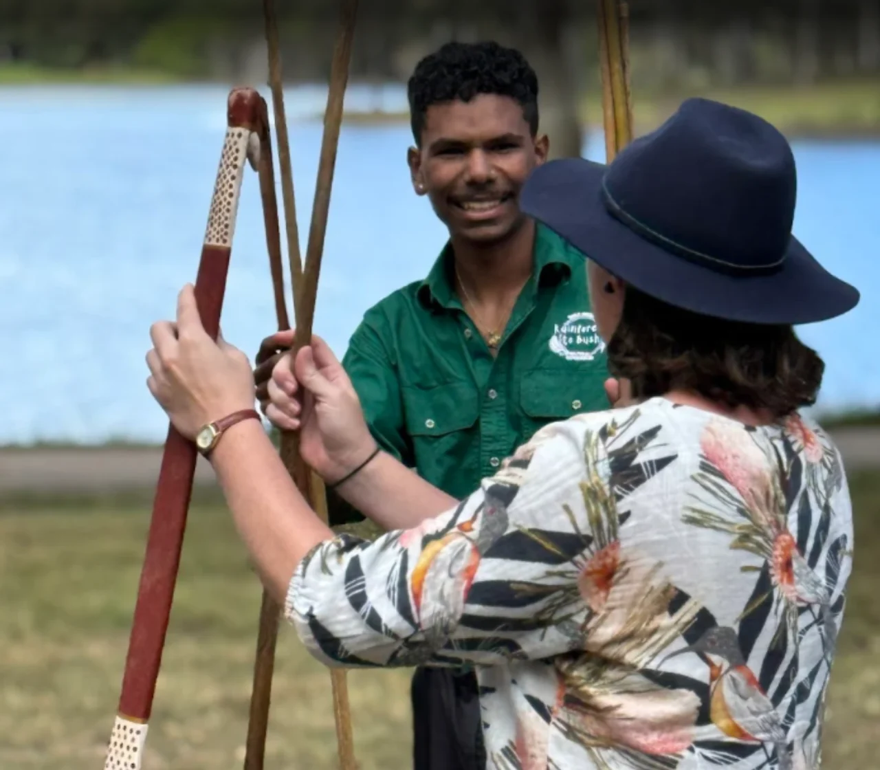 A young Indigenous guide supervises a visitor handling a decorated throwing implement and spear shafts beside water