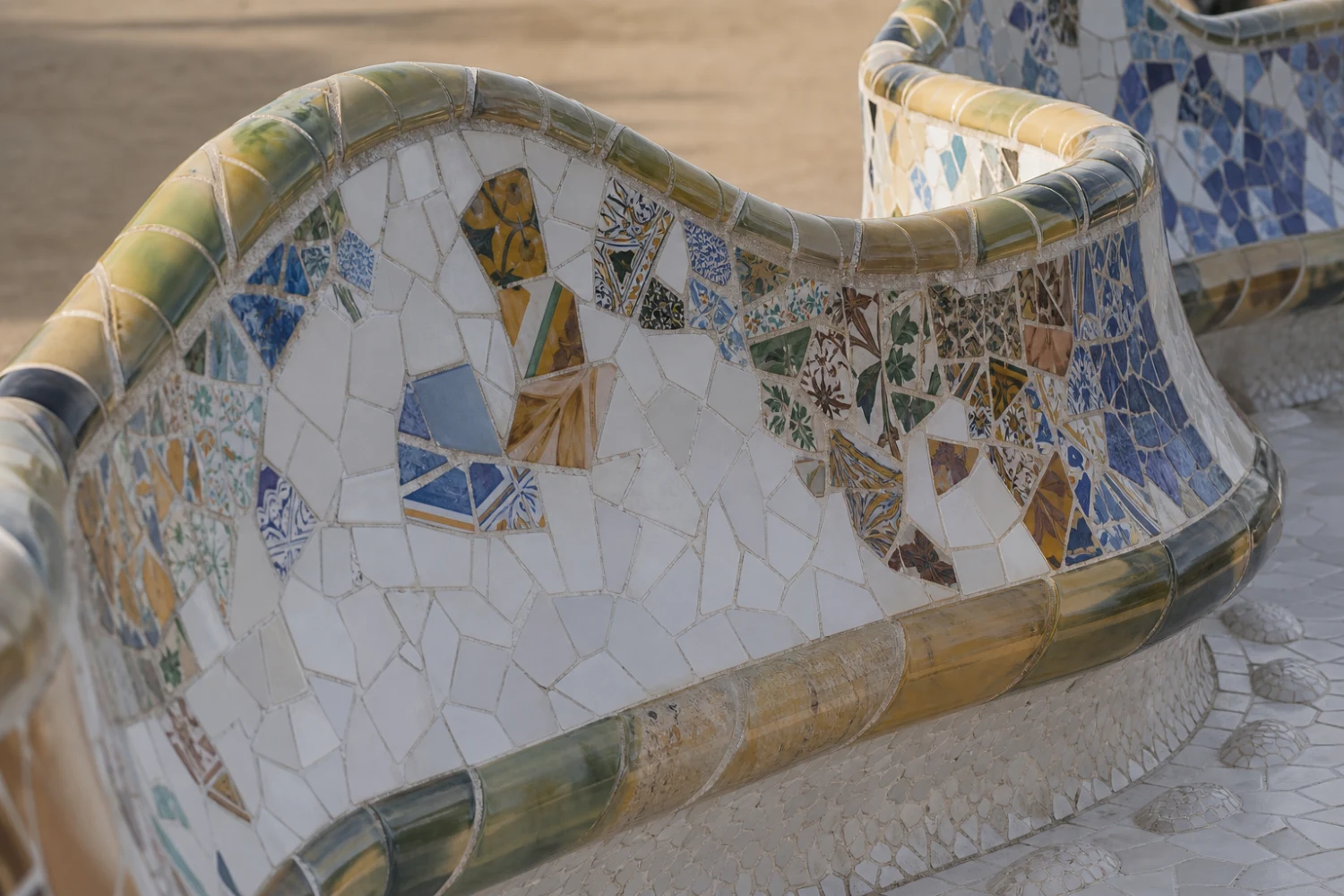 A close, raking view shows how irregular ceramic fragments wrap the bench’s compound curve and form colour fields