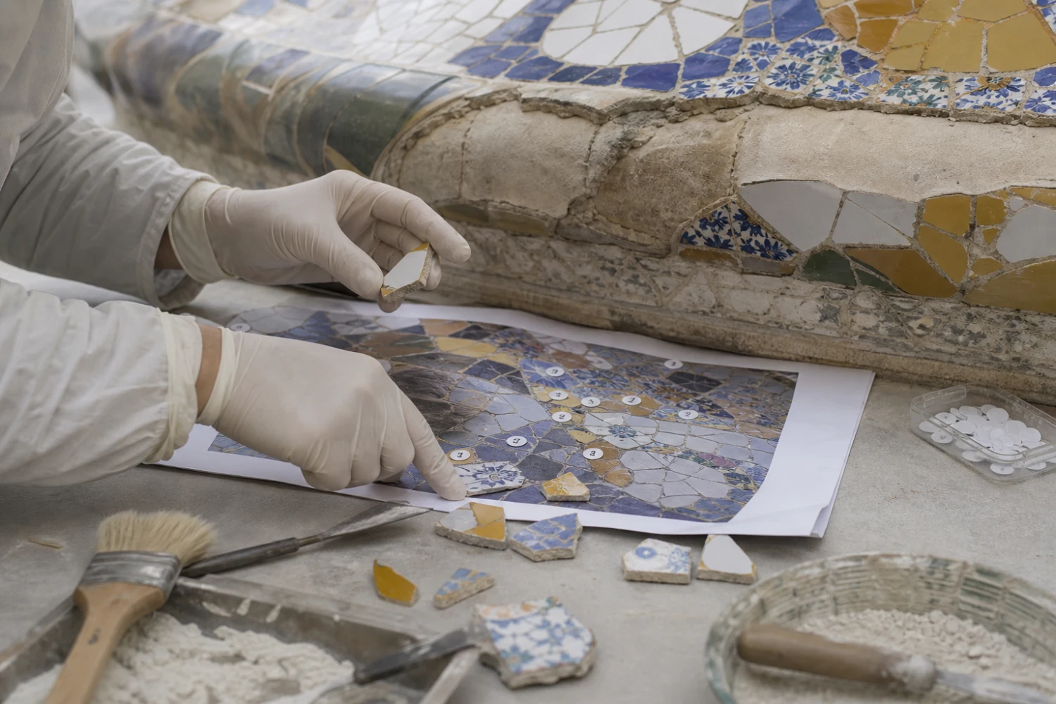 A conservator’s gloved hands document and reset trencadís fragments against a mapped ceramic surface