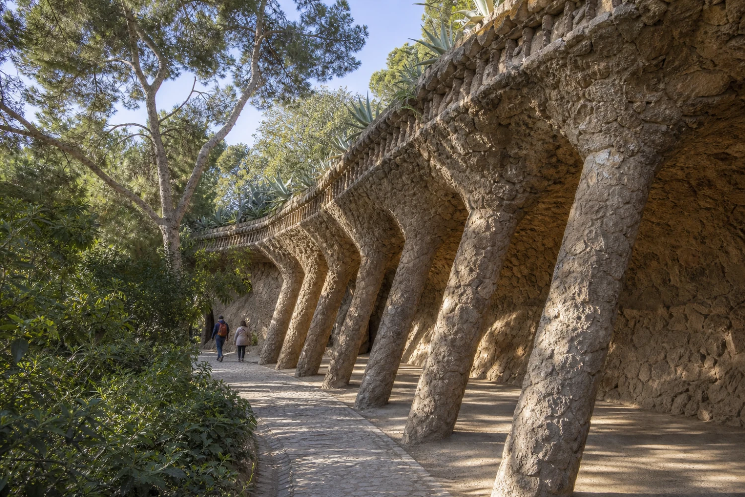 A Park Güell viaduct curves through planted ground on inclined columns of rough local stone