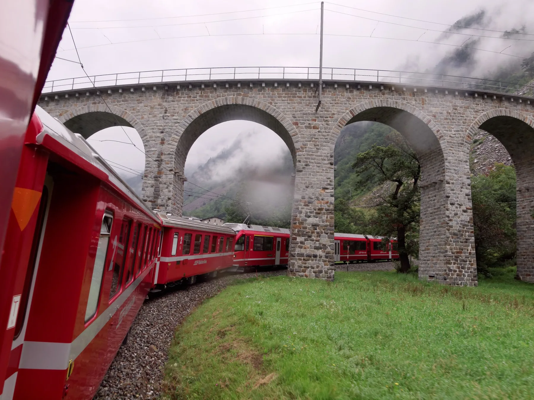 A red RhB train curves beneath the stone arches of the Brusio spiral viaduct in mist.
