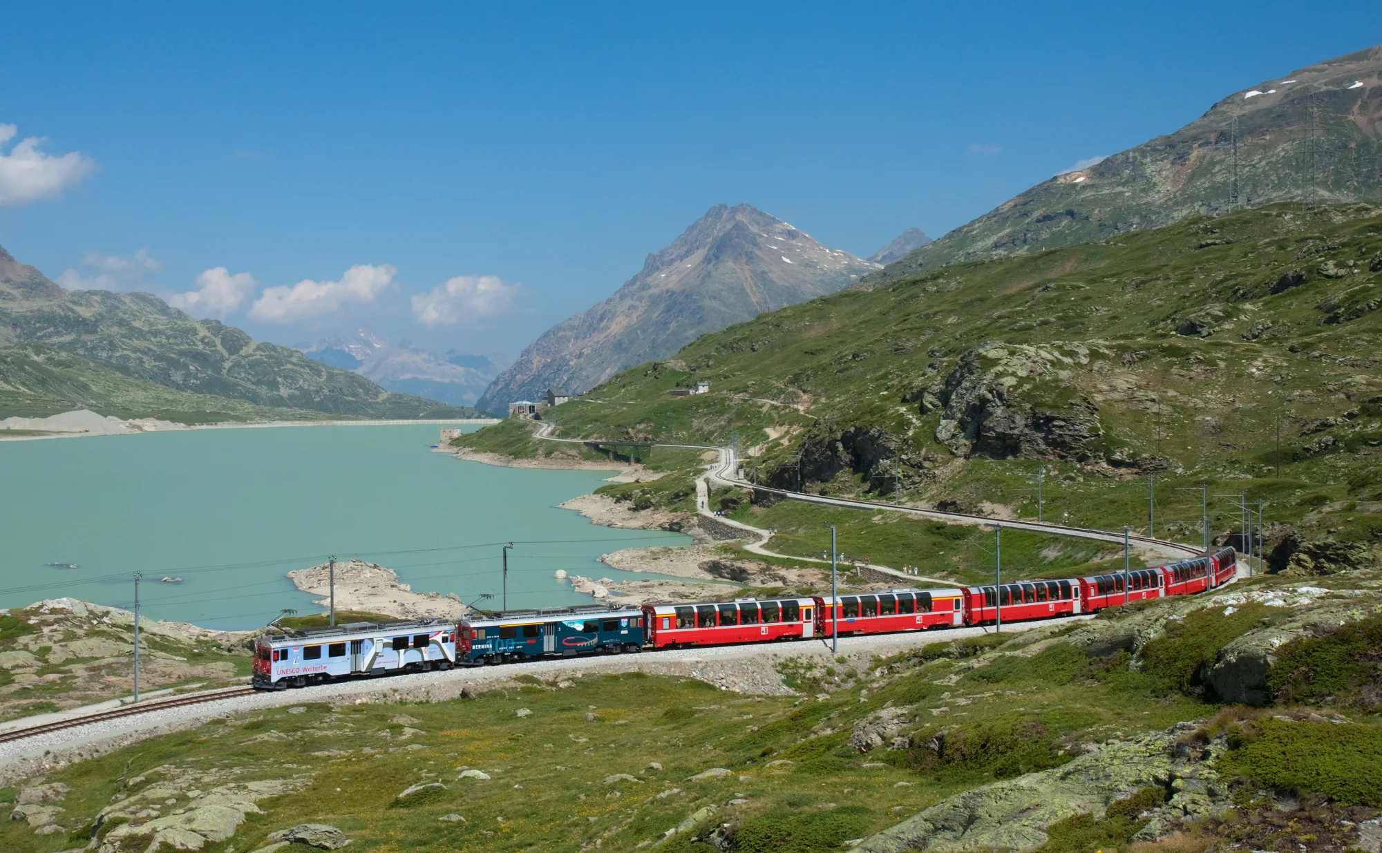 A red Rhaetian Railway train follows Lago Bianco below the Bernina massif