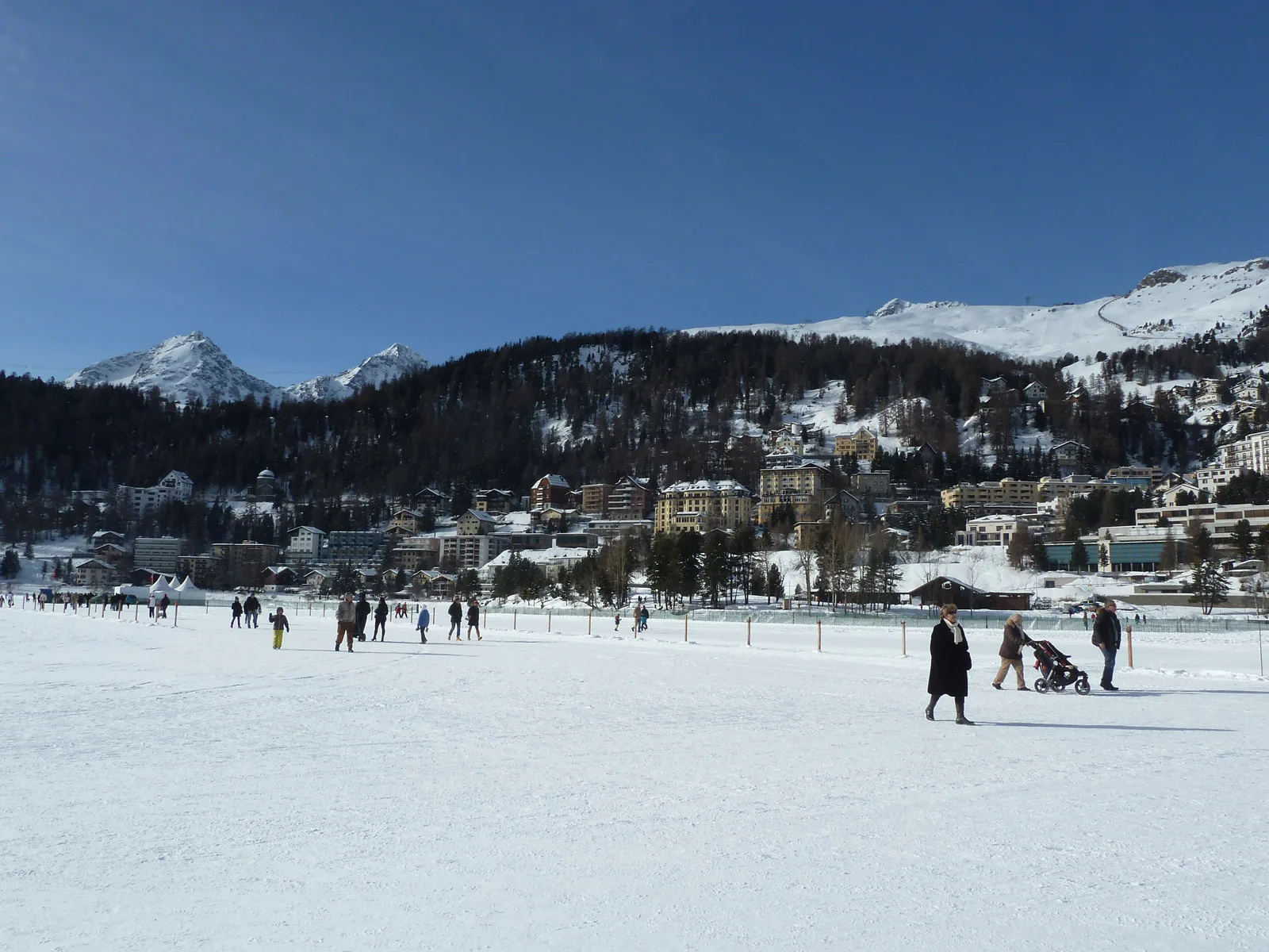 St. Moritz rises behind its frozen lake during winter activity, with hotel and residential buildings spreading across the slope.