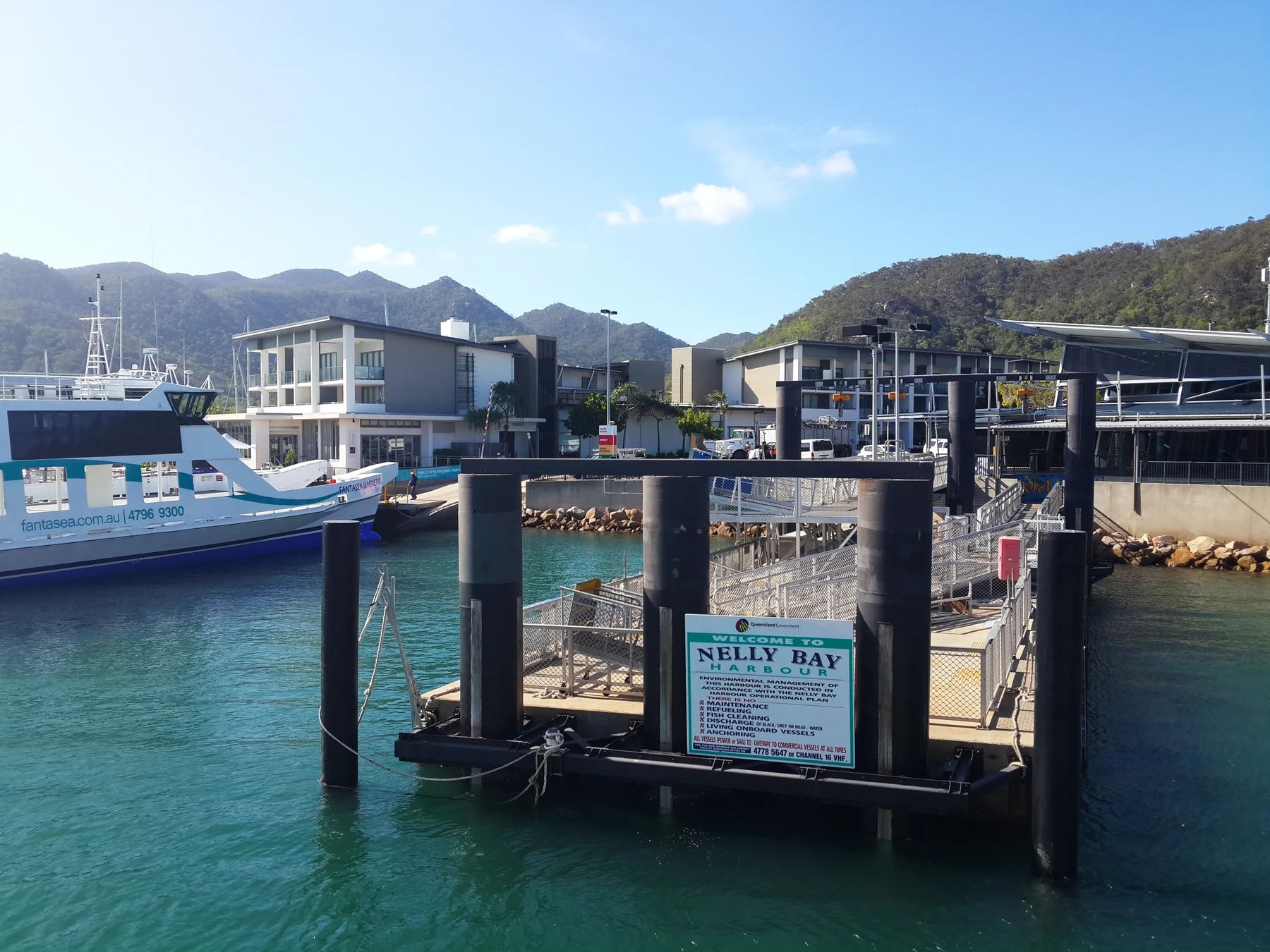 A historical documentary photograph shows the ferry-terminal building, pontoons and gangways at Nelly Bay beneath Yunbenun’s granite hills.