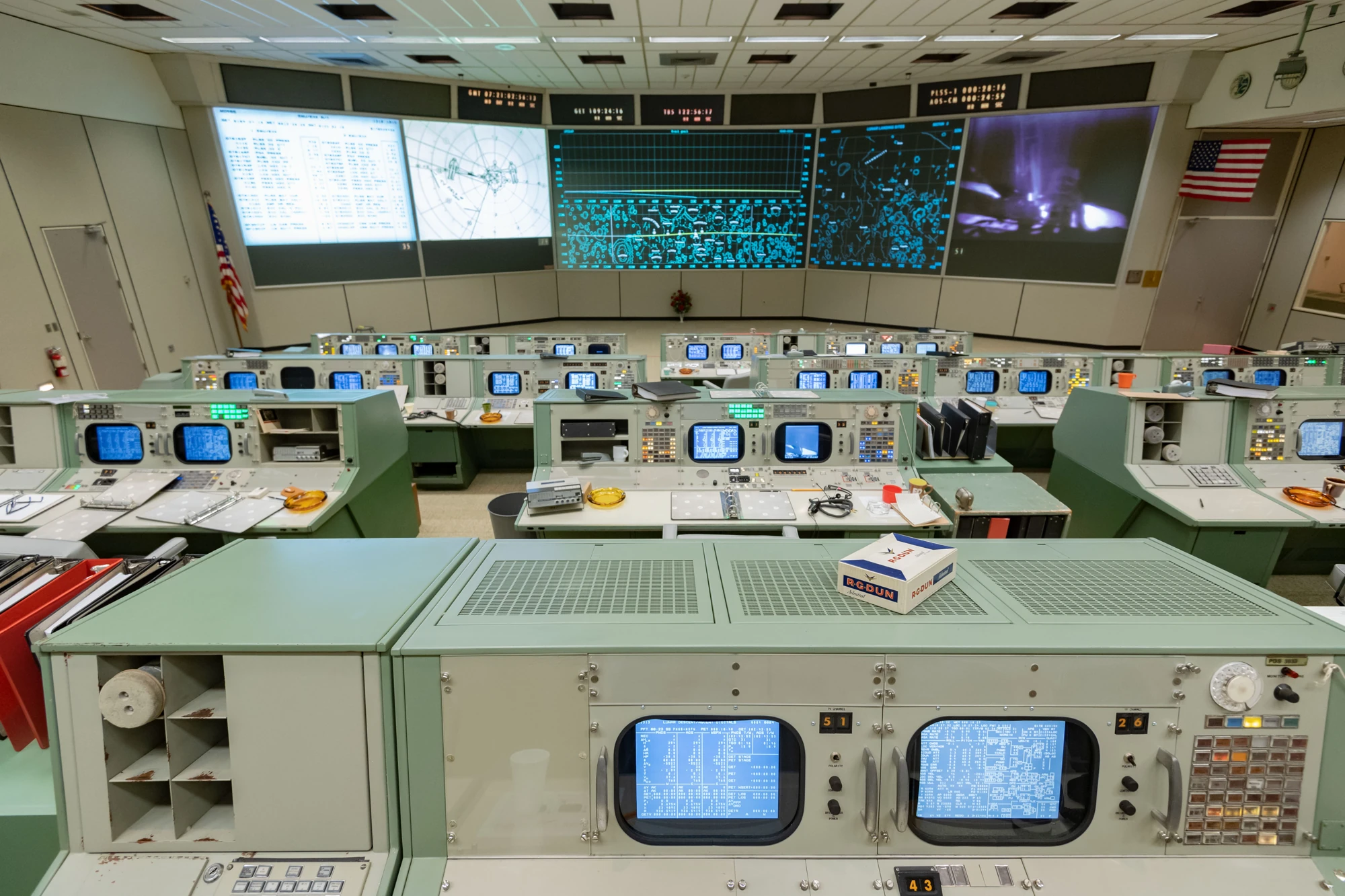Rows of restored Apollo Mission Control consoles with documents and personal objects, facing the front display screens