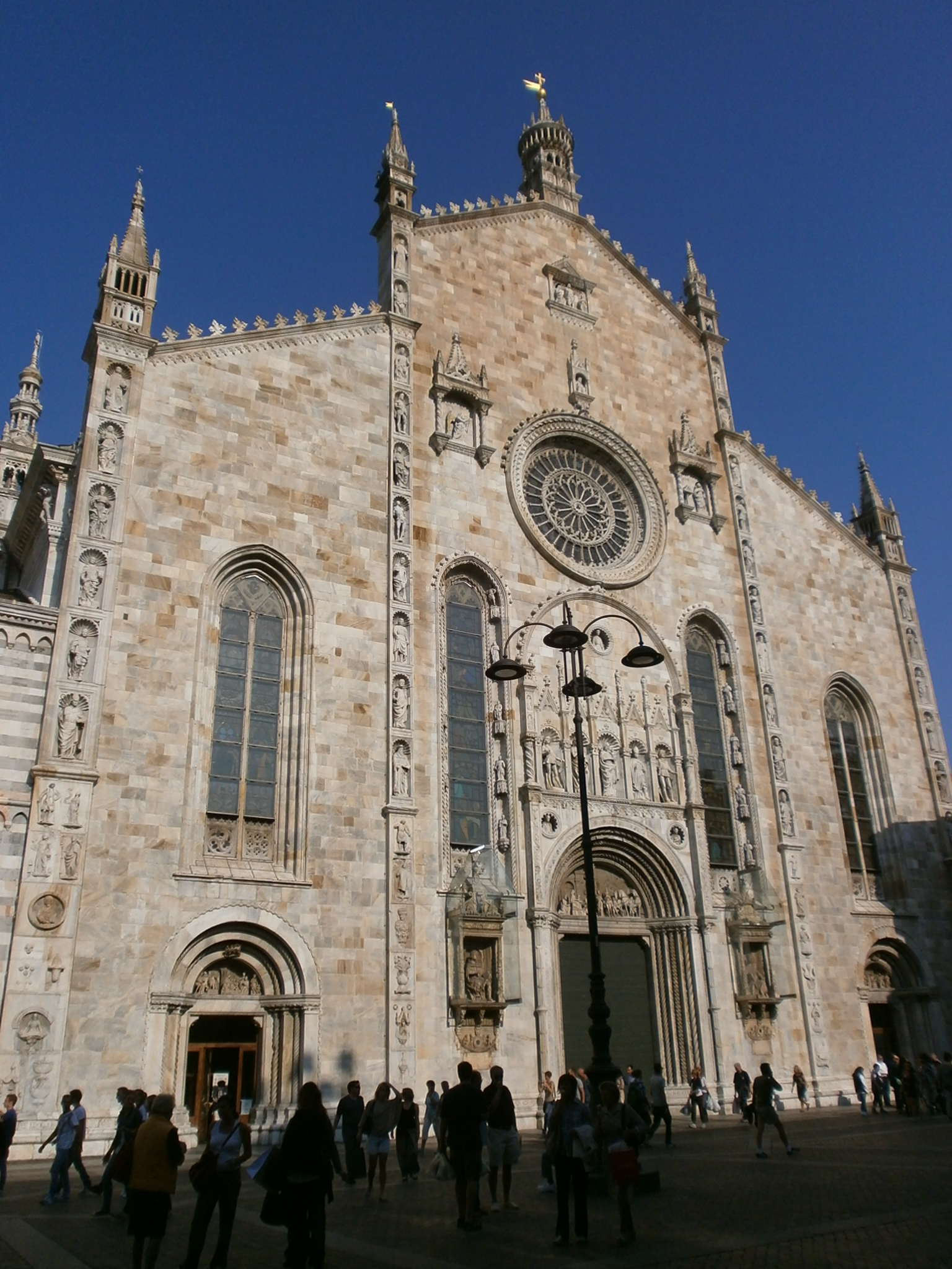 The pale stone façade of Como Cathedral rises above the piazza, with the rose window, sculpted portals and vertical figure bands visible