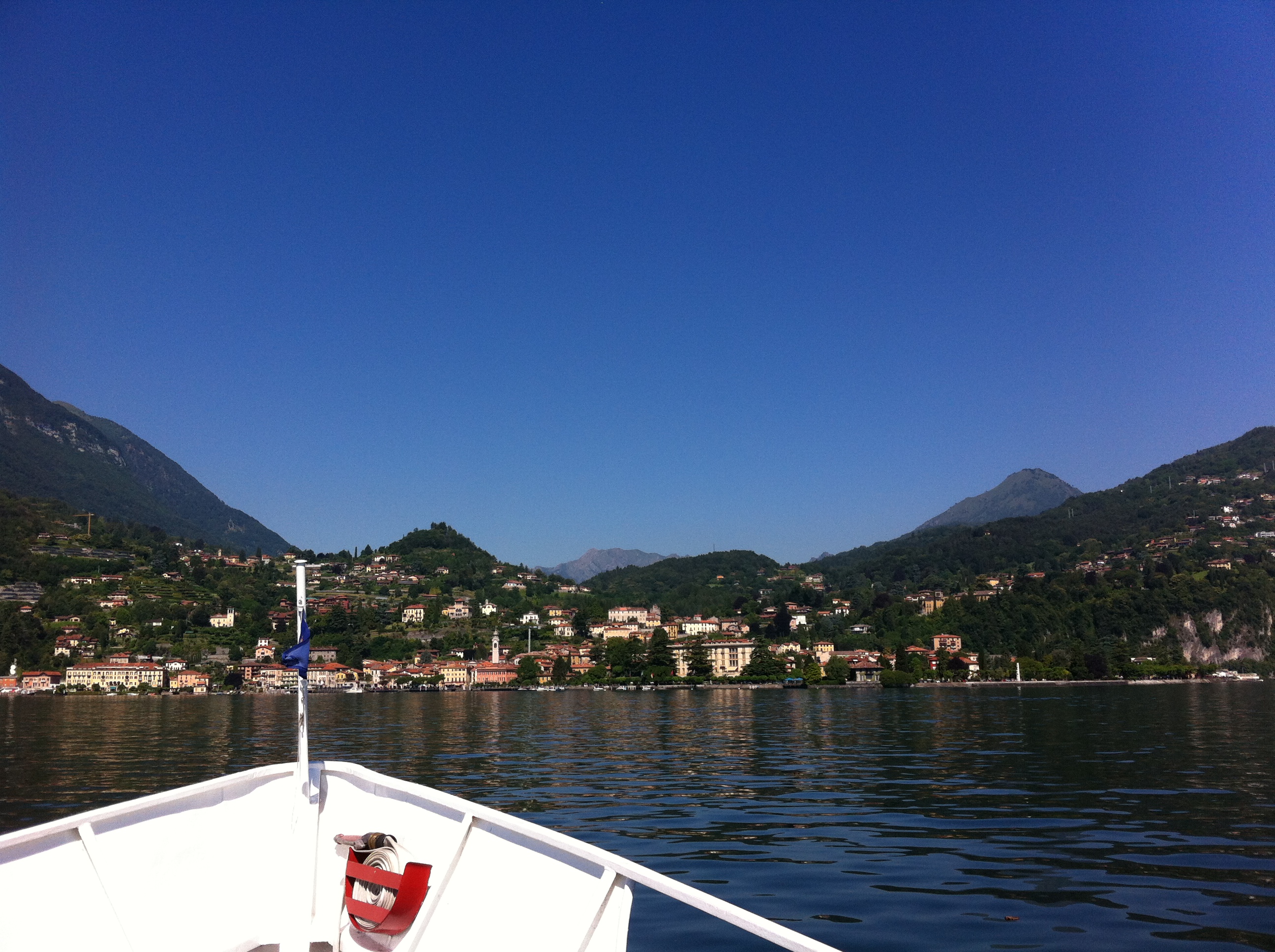 The bow of a passenger vessel points toward a Lake Como shore where houses and cultivated terraces occupy a narrow strip below steep hills
