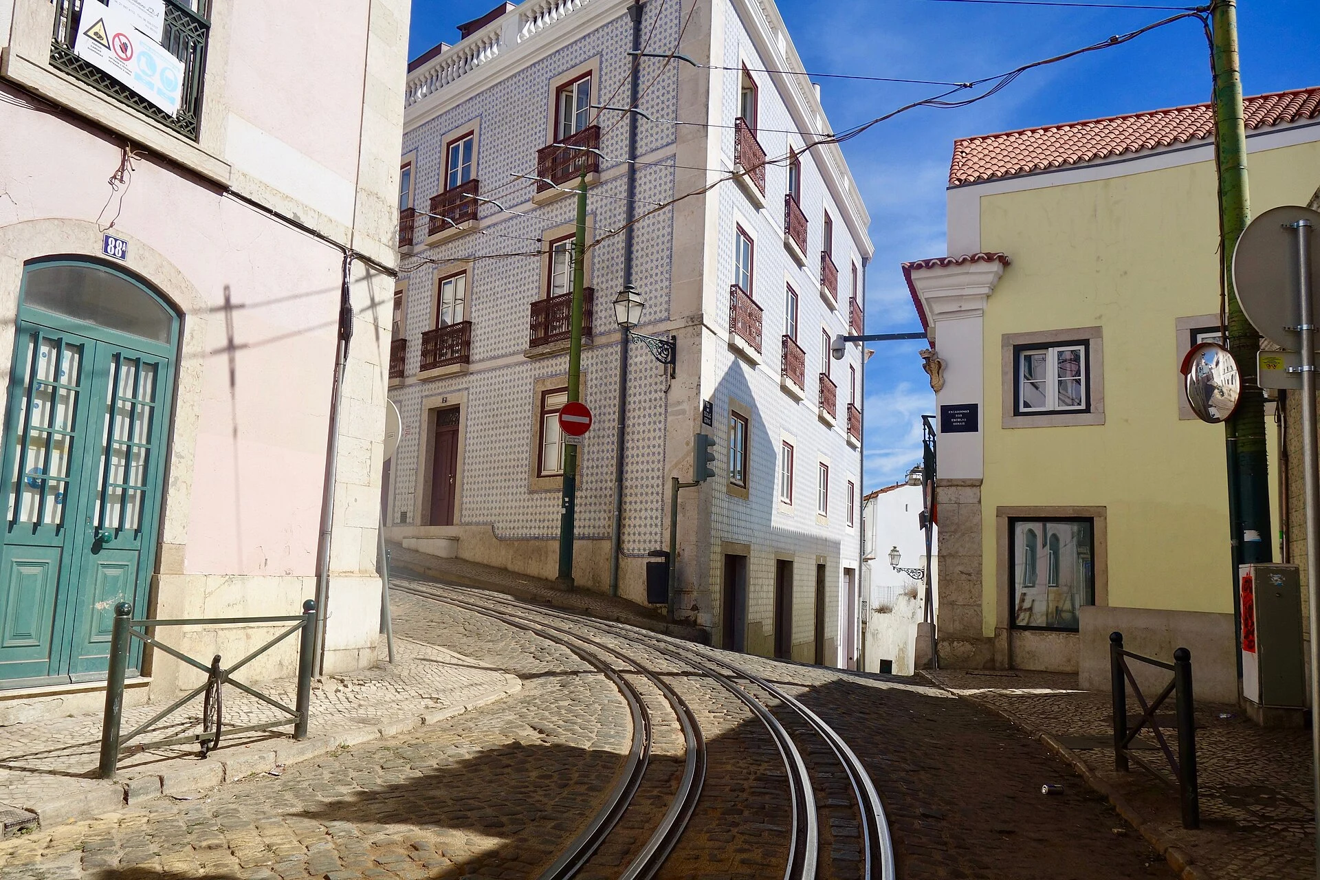 Tram rails curve through a steep Alfama street between narrow pavements, tiled façades and fixed street furniture.