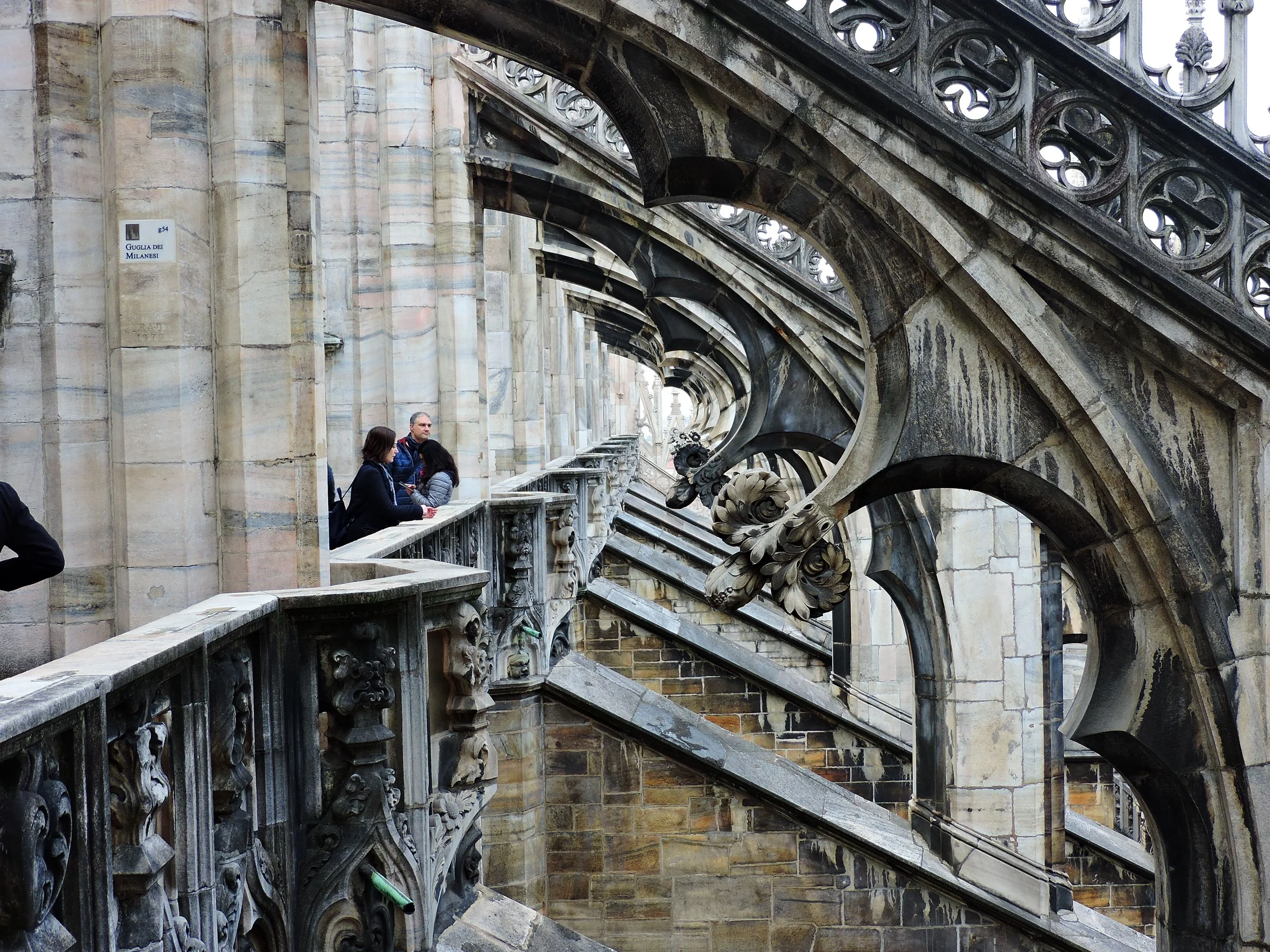 A terrace passage runs beside massive weathered arches, tracery, sculpted drainage and pale Candoglia marble