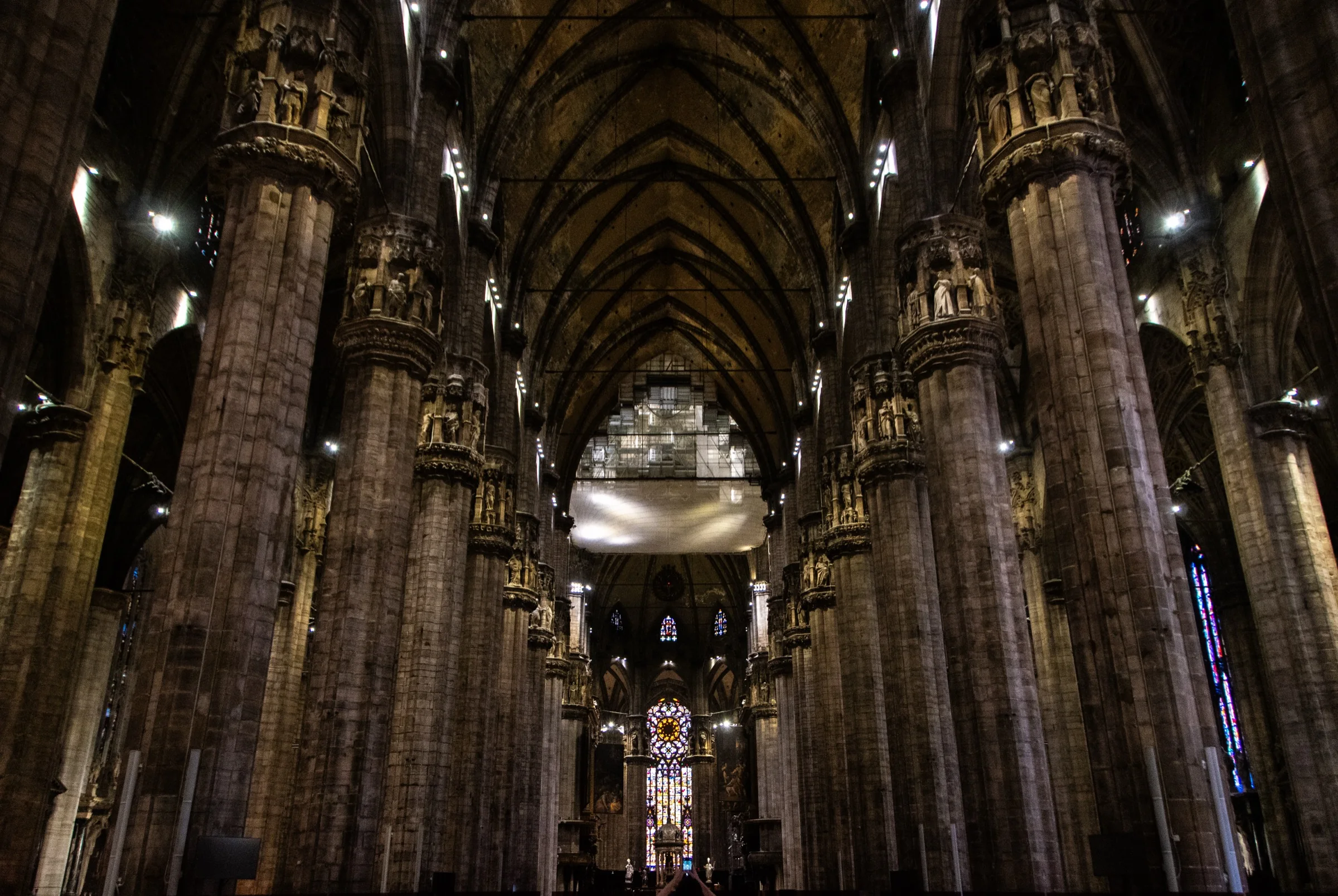 The dark central nave of Milan Cathedral with clustered piers, sculptural capitals, ribbed vaults and stained glass at the east end