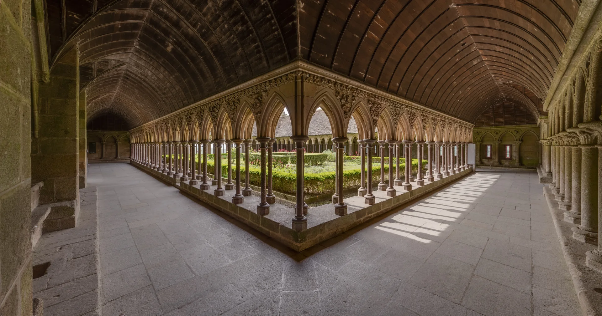 The Gothic cloister forms a luminous quadrangle of slim double columns around a garden, with timber vaulting above the covered walk.