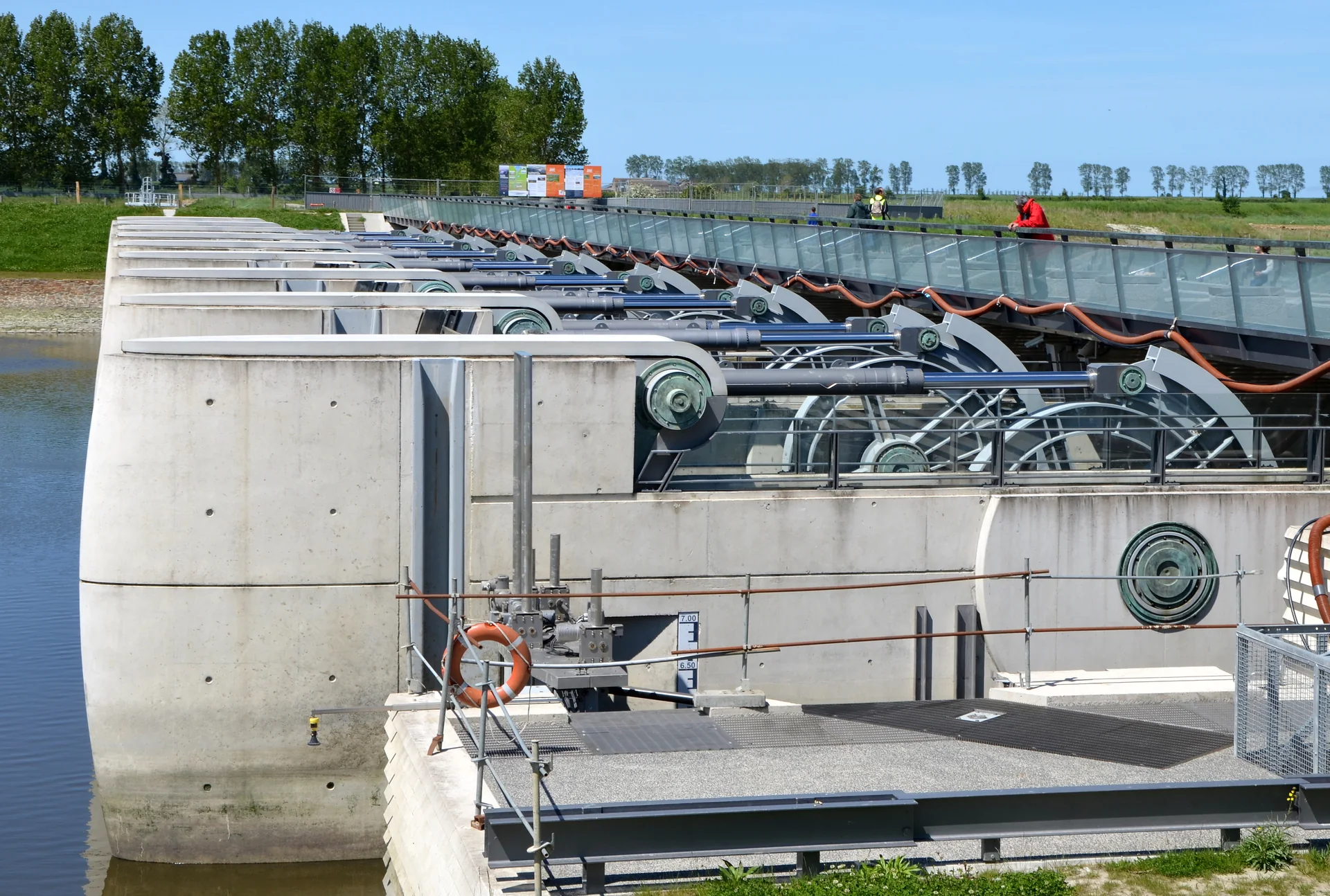 The Couesnon dam is seen close-up, with concrete bays, mechanical gates and the pedestrian deck crossing above the water-control structure.