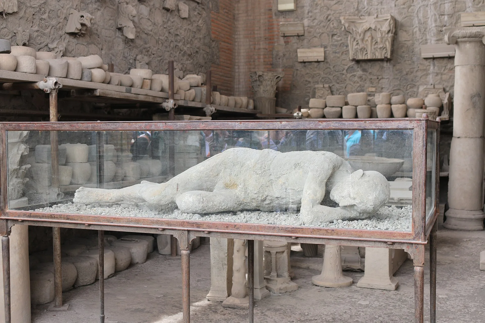 A single Pompeii body cast is displayed inside a glass case in the Forum Granary, with stored amphorae and architectural fragments behind it