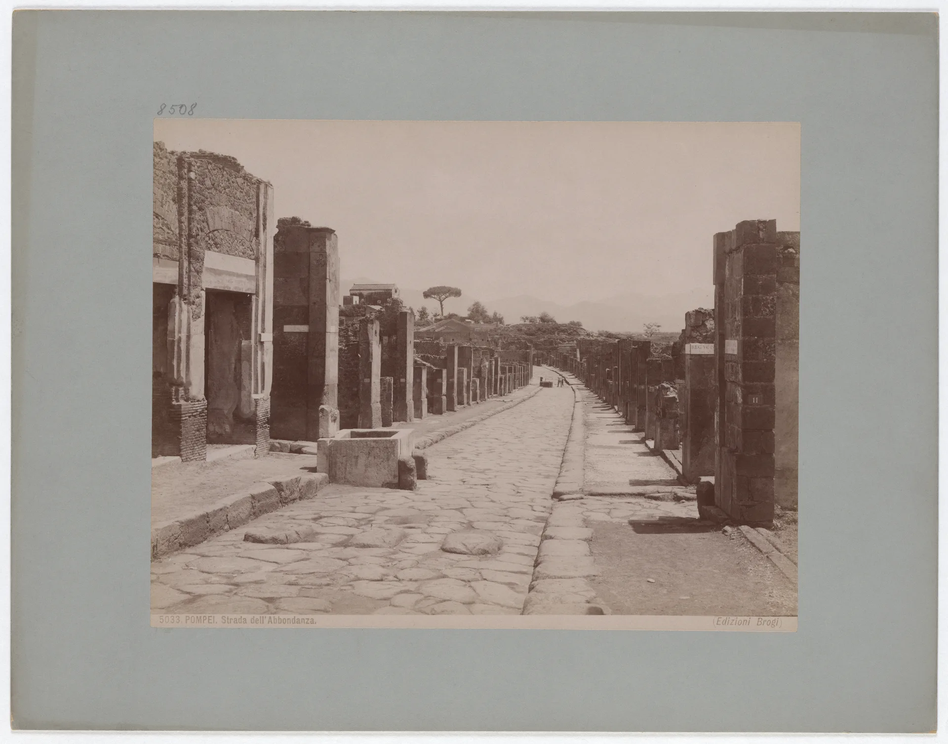 A nineteenth-century archival photograph looks down the excavated Via dell'Abbondanza at Pompeii, with doorways, fountains and high pavements