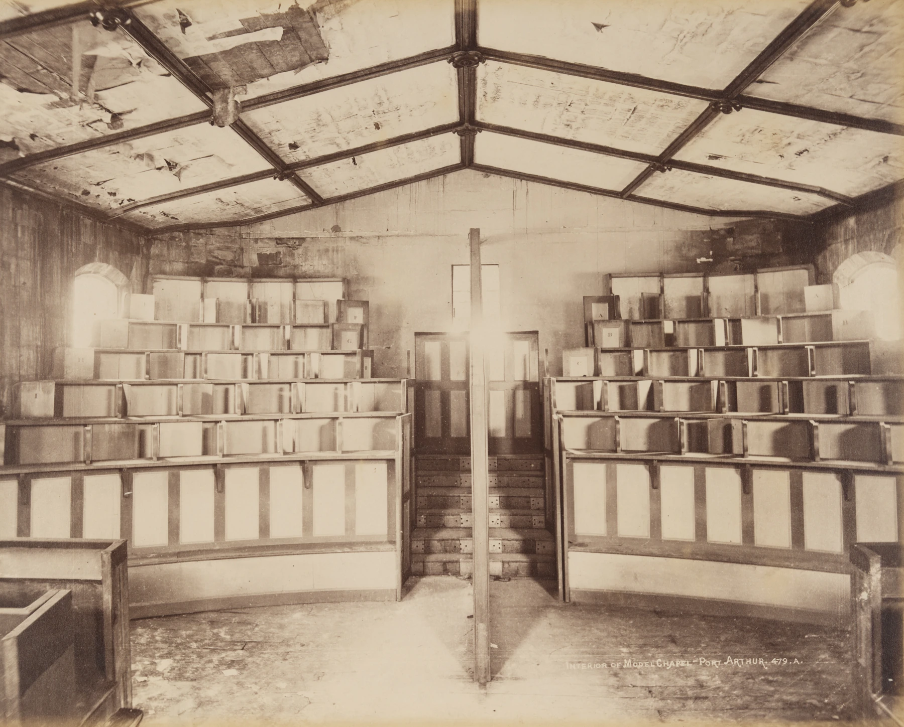 Individual wooden stalls rise in stepped rows inside the Model Chapel of the Separate Prison in an Anson Brothers archival photograph.