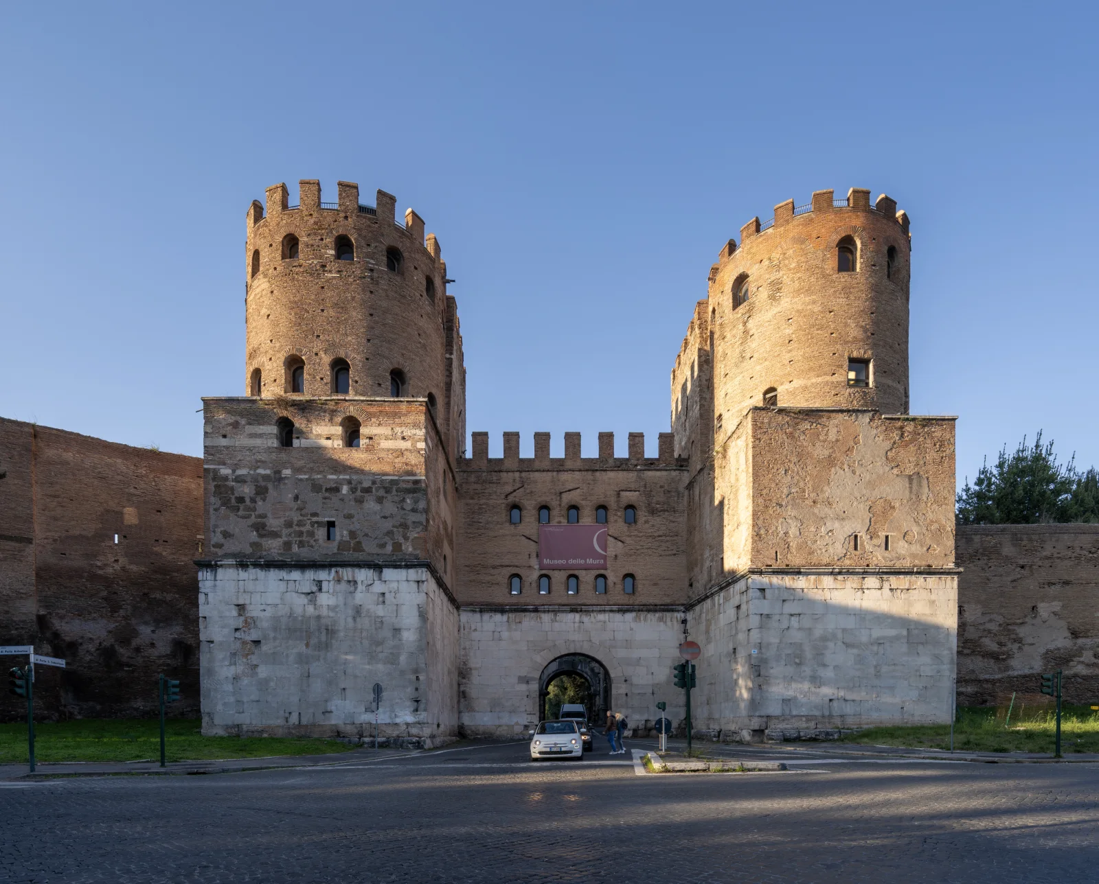 The twin towers and central opening of Porta San Sebastiano stand above the modern road.