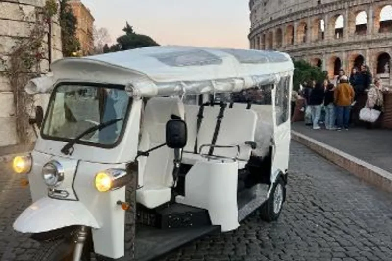The exact connected product's white electric tuk tuk stands beside the Colosseum, showing its compact three-wheel format and passenger bench.