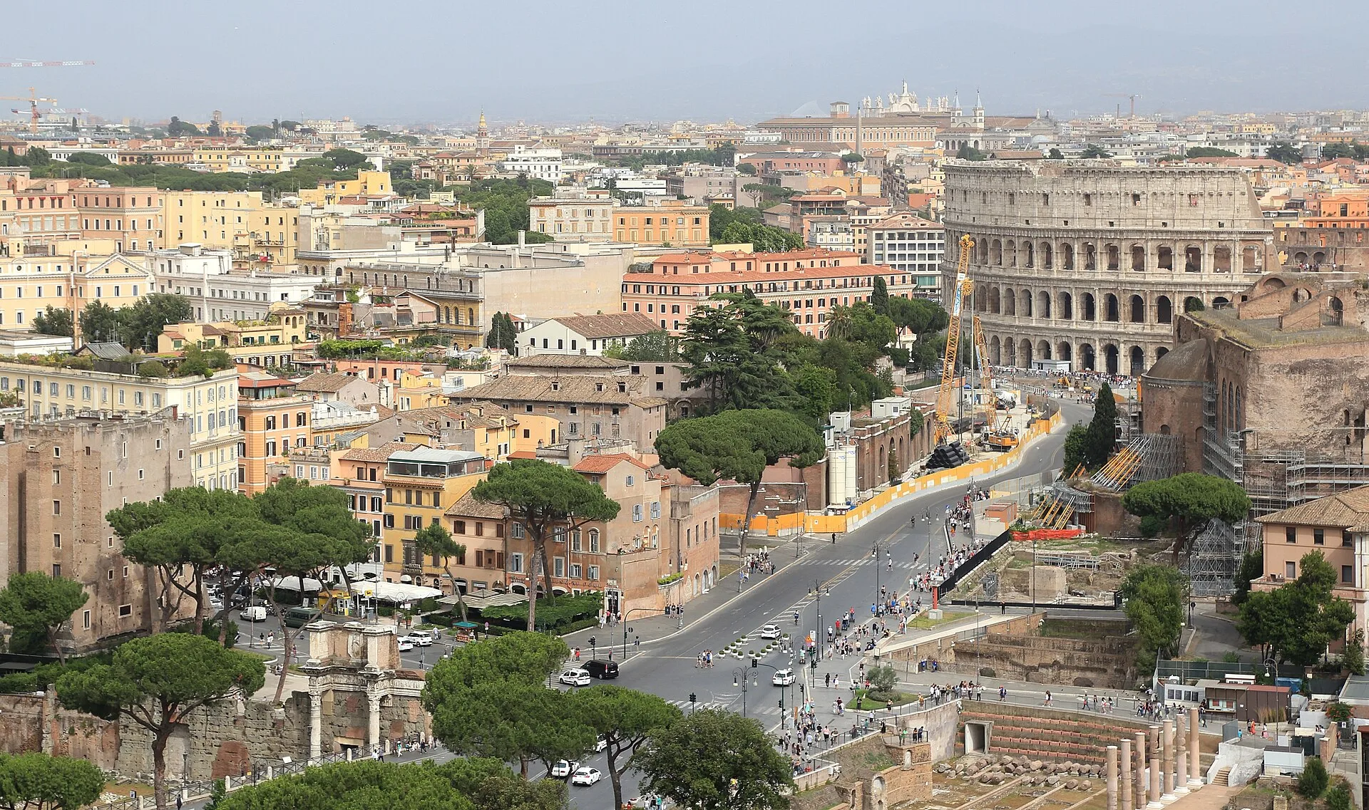 A high view along Via dei Fori Imperiali shows roadway, archaeological remains, later buildings and the Colosseum sharing one urban corridor.