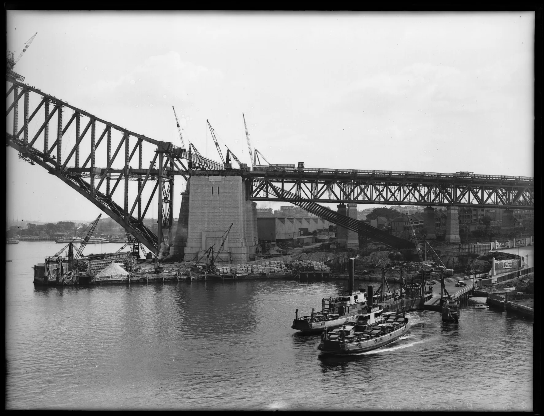 The Sydney Harbour Bridge under construction around 1930, with the arch extending over a working harbour and ferry traffic below.