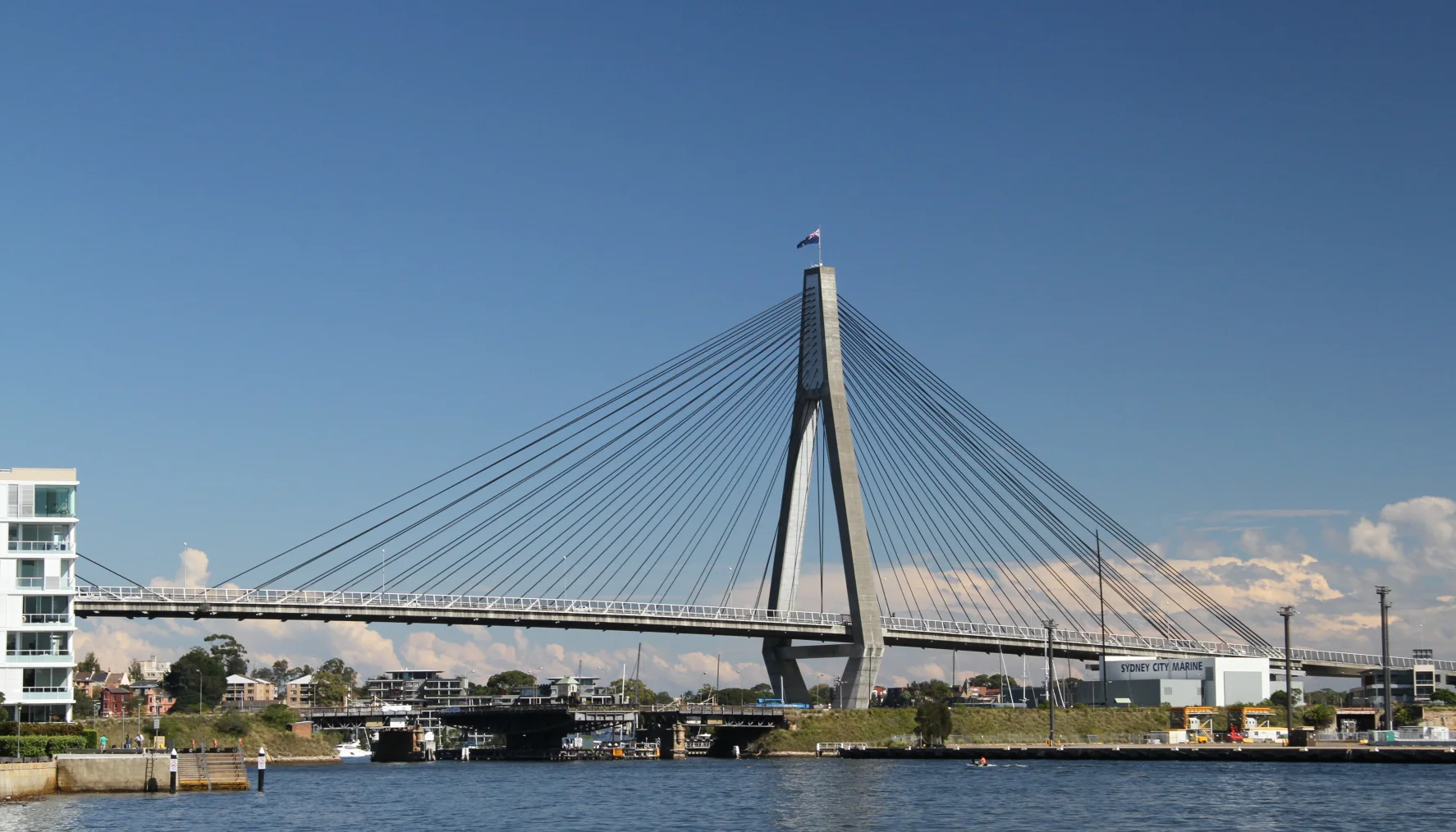 The ANZAC Bridge and maritime edge seen from Pirrama Park, with engineered shore, working-water traces and residential redevelopment sharing the frame.